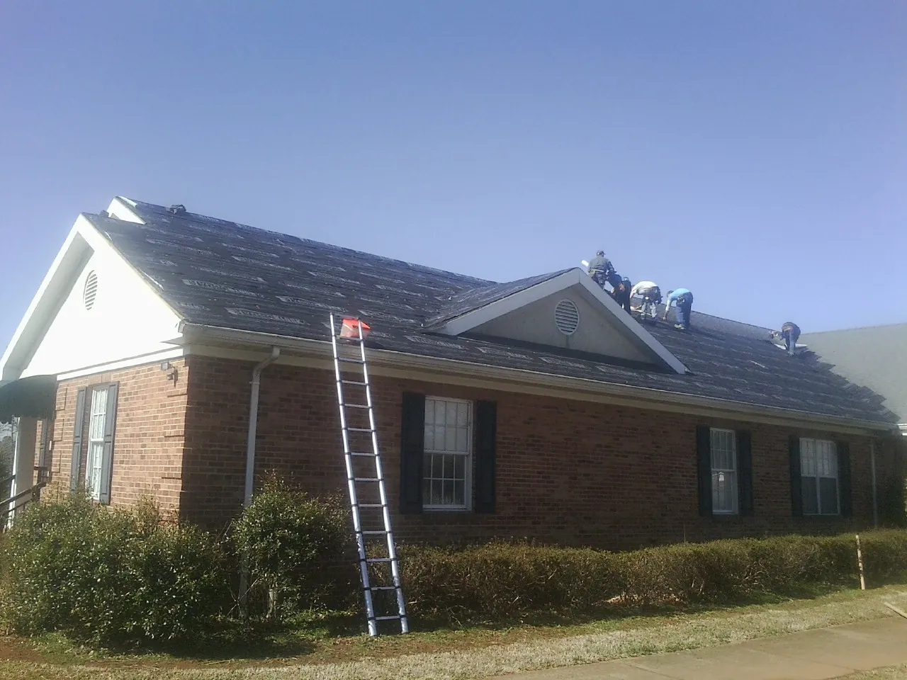 Crew tearing shingles off a brick ranch roof with a ladder set — Emergency Roof Repair work by our crew in Coker