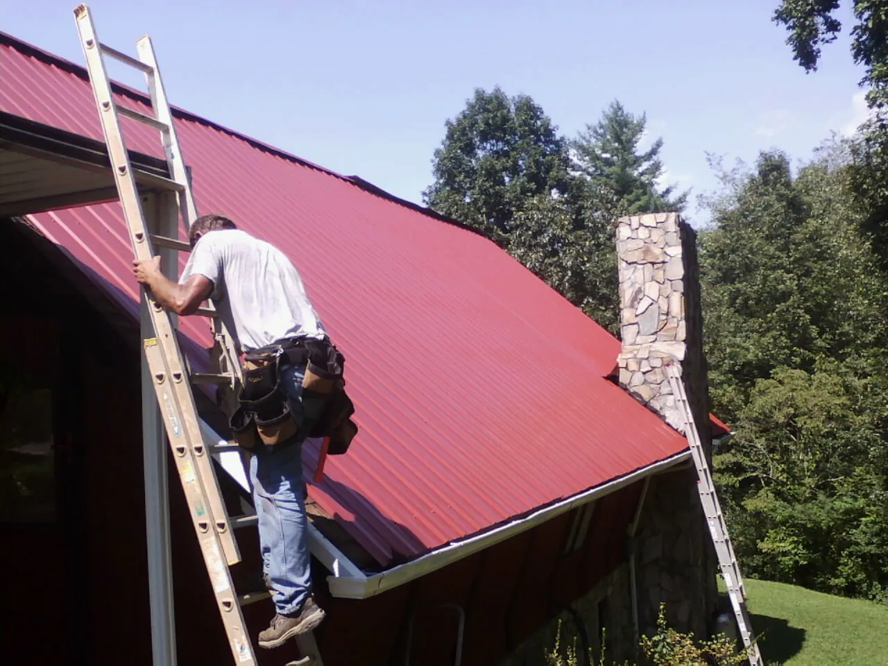 Roofer on a ladder beside a red metal roof and stone chimney — Soffit & Fascia Repair by Roofing Brothers in Coker